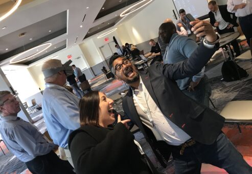 Two participants enthusiastically taking a selfie during The Infinite Loop team building event, while others engage in VR challenges in the background.