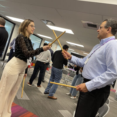 Two participants practicing rhythm coordination with drumsticks during a BeatsWork team building activity. thumbnail