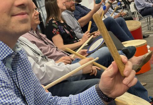 Participants seated in a circle, practicing rhythm on percussion instruments during a BeatsWork team building activity.
