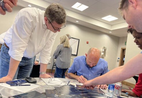Participants gather around a table during The Infinite Loop team building event, focusing on solving a collaborative puzzle with strategy sheets laid out in front of them.