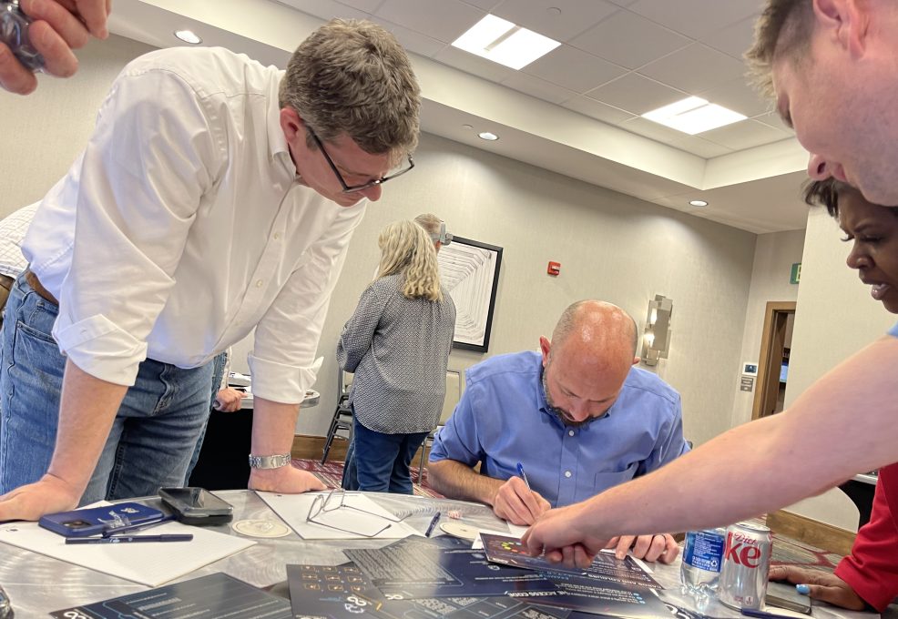 Participants gather around a table during The Infinite Loop team building event, focusing on solving a collaborative puzzle with strategy sheets laid out in front of them.