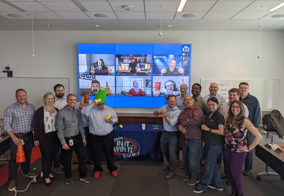 A group of participants posing together with props, smiling and enjoying the In It to Win It hybrid team building activity, with some team members visible on a large screen via video call, showcasing a blend of in-person and virtual participation.