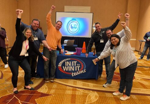 A group of participants celebrating with raised arms and smiling faces in front of a table labeled 'In It to Win It' during a team building activity, showcasing excitement, teamwork, and a fun, competitive atmosphere.