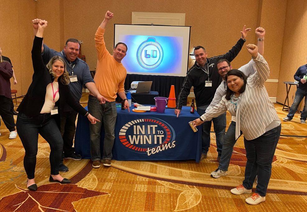 A group of participants celebrating with raised arms and smiling faces in front of a table labeled 'In It to Win It' during a team building activity, showcasing excitement, teamwork, and a fun, competitive atmosphere.
