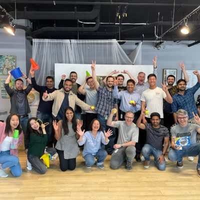 A large group of participants posing energetically with smiles, props, and raised hands, celebrating their participation in the In It to Win It team building event, showcasing a fun and lively group dynamic. thumbnail