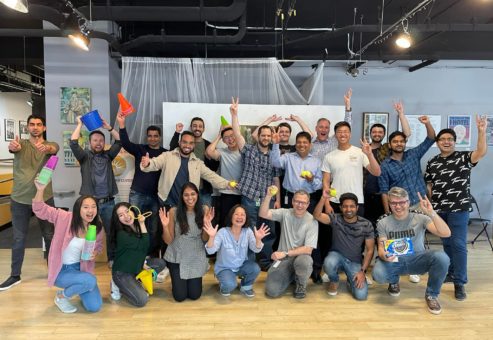 A large group of participants posing energetically with smiles, props, and raised hands, celebrating their participation in the In It to Win It team building event, showcasing a fun and lively group dynamic.