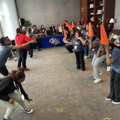 Participants engaged in a lively ring toss challenge, with some wearing traffic cones on their heads as targets, during the In It to Win It team building game, creating a fun and energetic atmosphere. thumbnail