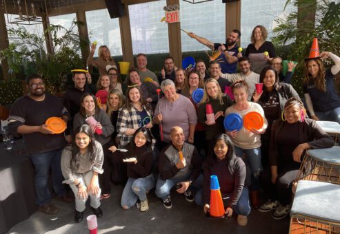 A large group of participants posing cheerfully with various props like cones, cups, frisbees, and rings, celebrating their participation in the In It to Win It team building event, showcasing a vibrant and fun team atmosphere.