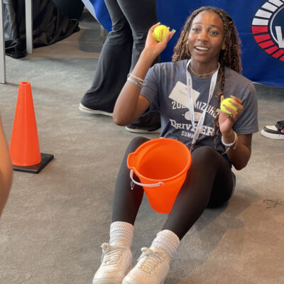 A participant sitting on the floor holding two tennis balls and an orange bucket, engaging in a challenge during the In It to Win It team building event, demonstrating focus, coordination, and a playful spirit. thumbnail