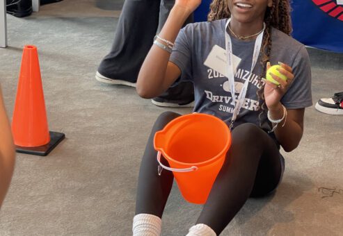 A participant sitting on the floor holding two tennis balls and an orange bucket, engaging in a challenge during the In It to Win It team building event, demonstrating focus, coordination, and a playful spirit.