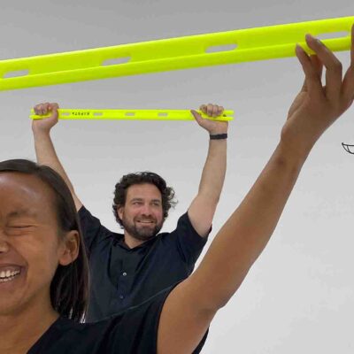 Participants lift obstacle bars with excitement as a drone flies by during the Game of Drones team building challenge, emphasizing fun and teamwork. thumbnail