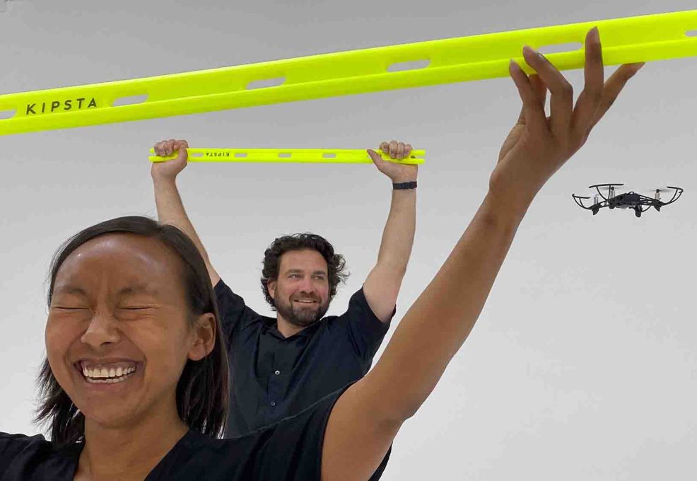 Participants lift obstacle bars with excitement as a drone flies by during the Game of Drones team building challenge, emphasizing fun and teamwork.