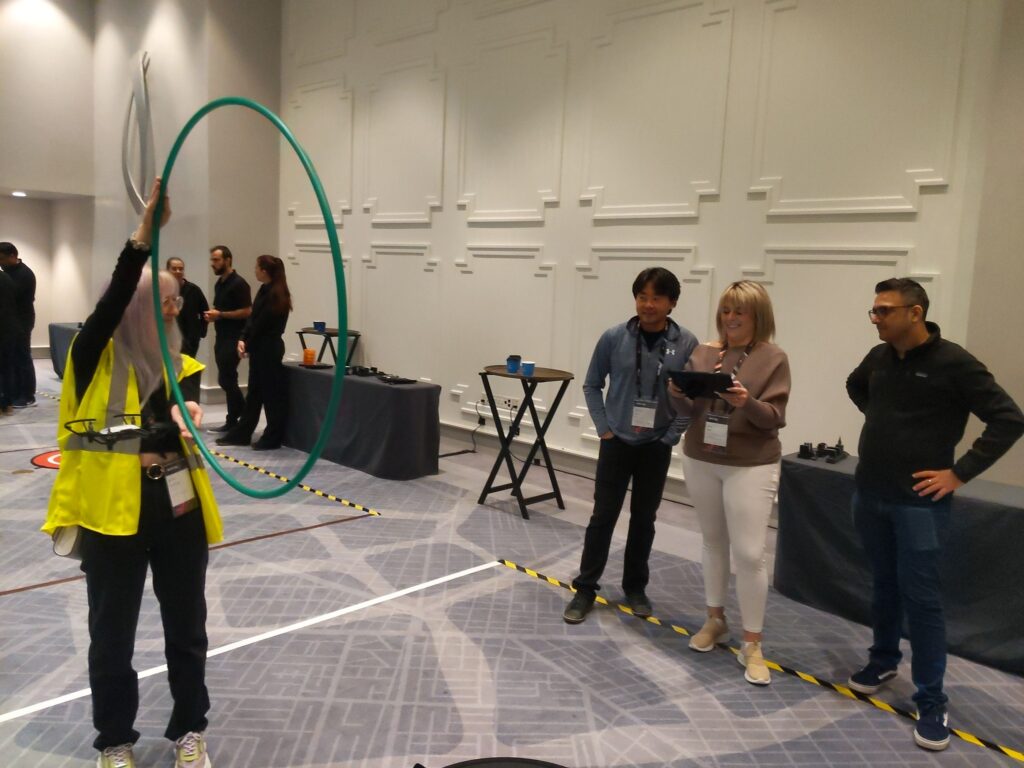 A participant guides a drone through a hula hoop obstacle while teammates monitor and strategize during the Game of Drones team building event.