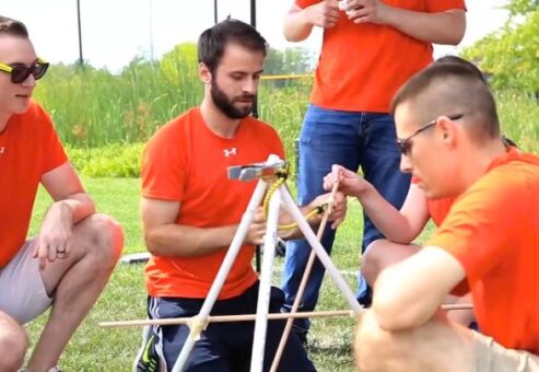 A team of four participants wearing matching orange shirts working together outdoors on constructing a wooden catapult during the Catapult to Success team building activity, highlighting teamwork, creativity, and collaboration.