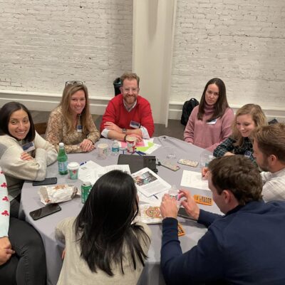 A group of participants sits around a table, smiling and engaged in conversation during the Helping Hands team building event. They are working together on tasks related to assembling care kits for the community. thumbnail