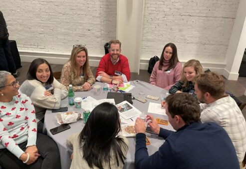 A group of participants sits around a table, smiling and engaged in conversation during the Helping Hands team building event. They are working together on tasks related to assembling care kits for the community.