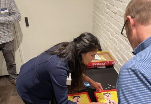 Participants engage in a fun challenge, playing the game 'Operation' as part of the Helping Hands team building event. The activity is designed to earn supplies for care kits through interactive and skill-based tasks.