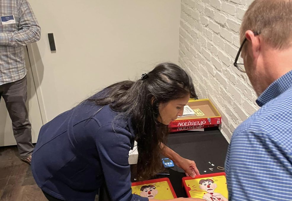 Participants engage in a fun challenge, playing the game 'Operation' as part of the Helping Hands team building event. The activity is designed to earn supplies for care kits through interactive and skill-based tasks.