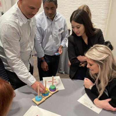 Participants gather around a table, engaged in a problem-solving activity involving colorful rings and pegs during the Helping Hands team building event. The group is focused and collaborating to complete the challenge. thumbnail