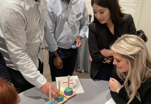 Participants gather around a table, engaged in a problem-solving activity involving colorful rings and pegs during the Helping Hands team building event. The group is focused and collaborating to complete the challenge.