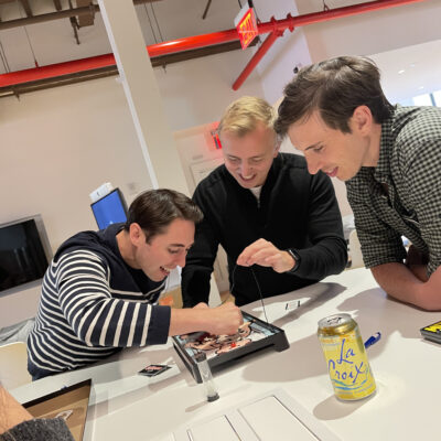 Three participants enthusiastically play the game 'Operation' at a table during the Helping Hands team building event. They are laughing and focused on completing the challenge to earn supplies for care kits. thumbnail