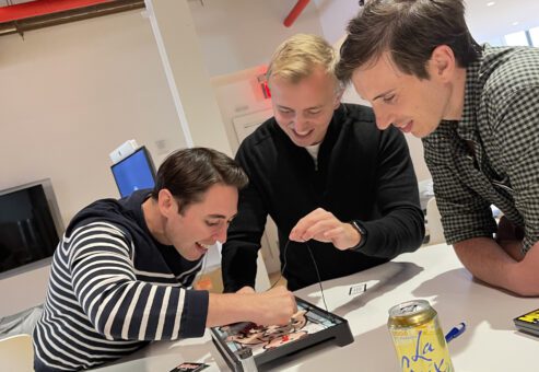Three participants enthusiastically play the game 'Operation' at a table during the Helping Hands team building event. They are laughing and focused on completing the challenge to earn supplies for care kits.