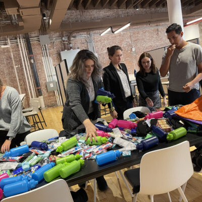 Participants gather around a table filled with supplies as they work together to assemble care kits during the Helping Hands team building event. The group organizes items such as water bottles, hygiene products, and snacks for community members in need. thumbnail