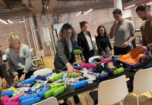 Participants gather around a table filled with supplies as they work together to assemble care kits during the Helping Hands team building event. The group organizes items such as water bottles, hygiene products, and snacks for community members in need.