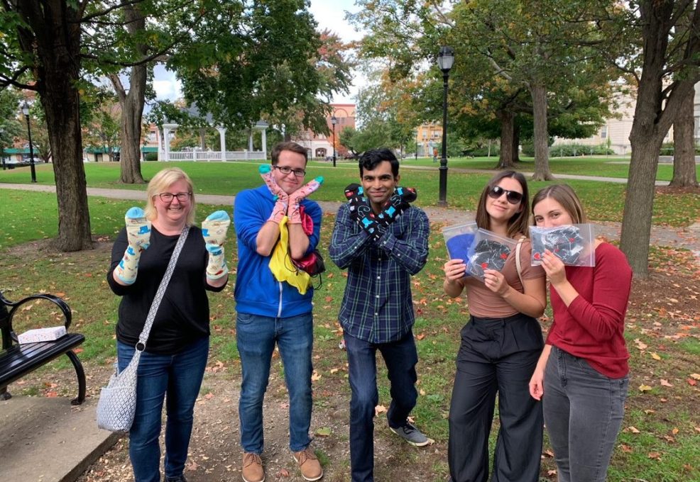 A group of five participants is standing outside in a park, each holding up socks and care items collected for a Healthcare Heroes scavenger hunt team building challenge.