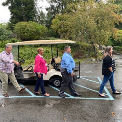 Team members recreate the iconic Abbey Road crossing in a lighthearted moment during the Healthcare Heroes team building event. Four participants walk in a line in front of a golf cart, mimicking the famous Beatles pose, bringing fun and engagement to the event which embraces Corporate Social Responsibility by supporting local healthcare professionals. thumbnail