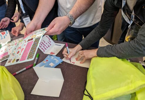 Participants are seen decorating cards with stickers and writing messages for healthcare professionals as part of the Healthcare Heroes scavenger hunt team building event. Bright green donation bags are on the table, ready to be filled with appreciation items.