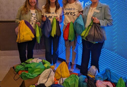 Four participants smiling and holding colorful drawstring bags filled with supplies, standing next to a large pile of completed donation bags as a result of a successful Healtchare Heroes team building scavenger hunt.