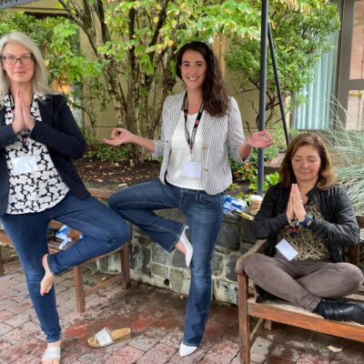Three team members engage in a relaxing yoga pose outdoors during the Healthcare Heroes team building event. Two participants stand in tree pose while the third sits cross-legged on a bench with her hands in prayer, representing mindfulness and relaxation. This interactive experience embraces Corporate Social Responsibility, helping teams collect items for donation to local hospitals. thumbnail