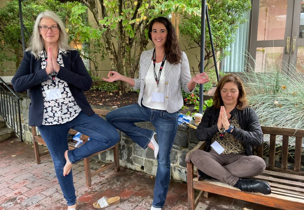Three team members engage in a relaxing yoga pose outdoors during the Healthcare Heroes team building event. Two participants stand in tree pose while the third sits cross-legged on a bench with her hands in prayer, representing mindfulness and relaxation. This interactive experience embraces Corporate Social Responsibility, helping teams collect items for donation to local hospitals.