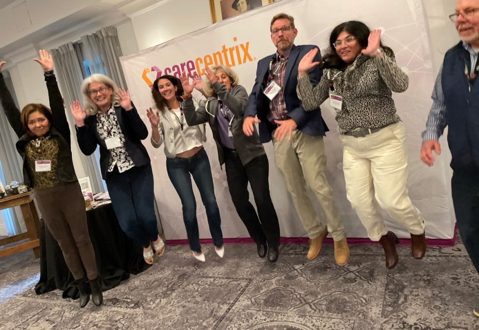 A group of smiling team members enthusiastically jump in the air in front of a CareCentrix banner, celebrating their team building event. They are participating in the Healthcare Heroes program, an interactive team building activity centered on Corporate Social Responsibility, designed to appreciate healthcare workers by assembling donation bags for local hospitals.