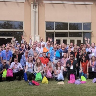 A large group of participants standing outdoors, many holding colorful drawstring bags, gathered in front of a building with tall windows. They are celebrating the accomplishment of a successful Healthcare Heroes team building scavenger hunt. thumbnail