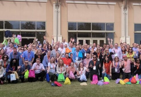 A large group of participants standing outdoors, many holding colorful drawstring bags, gathered in front of a building with tall windows. They are celebrating the accomplishment of a successful Healthcare Heroes team building scavenger hunt.