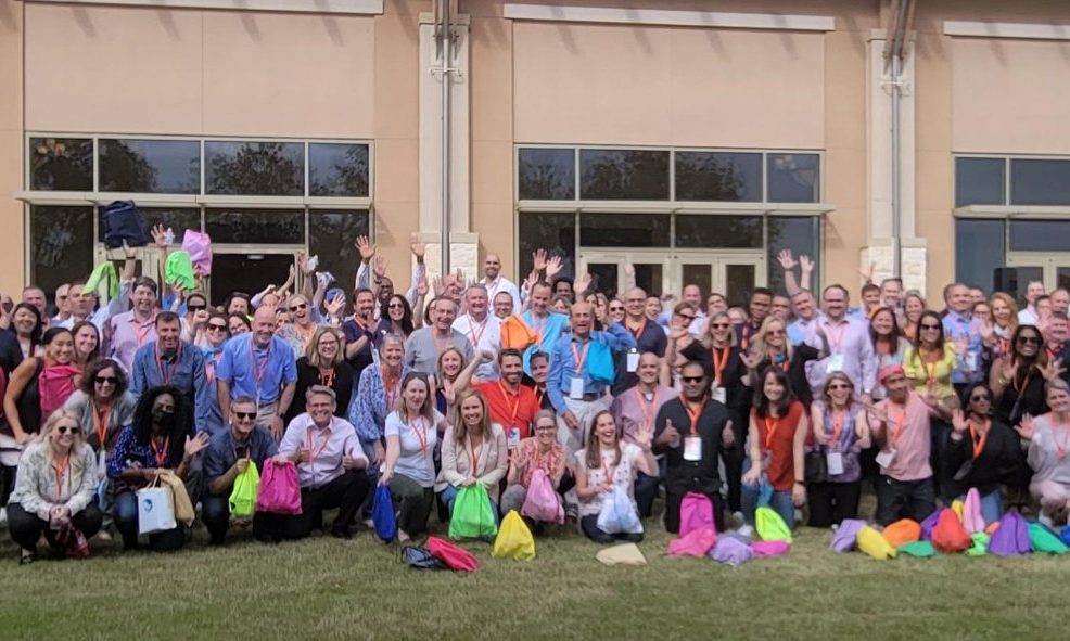 A large group of participants standing outdoors, many holding colorful drawstring bags, gathered in front of a building with tall windows. They are celebrating the accomplishment of a successful Healthcare Heroes team building scavenger hunt.