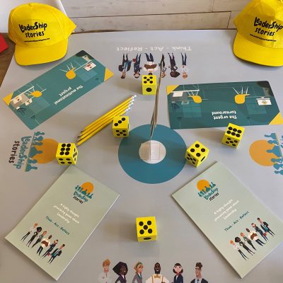 Table setup for the Leadership Stories game, featuring game materials like yellow dice, pencils, booklets, and colorful hats, designed for an interactive leadership team building experience. thumbnail