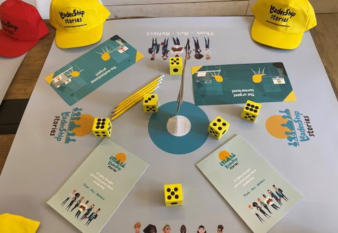 Table setup for the Leadership Stories game, featuring game materials like yellow dice, pencils, booklets, and colorful hats, designed for an interactive leadership team building experience.