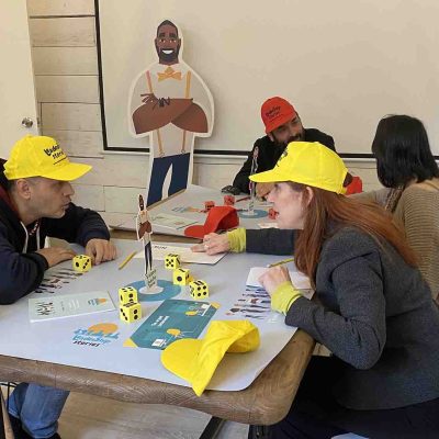 Team members engaging in the Leadership Stories game, wearing yellow hats and playing with game materials like dice and figurines, in a team building environment designed to enhance leadership skills. thumbnail