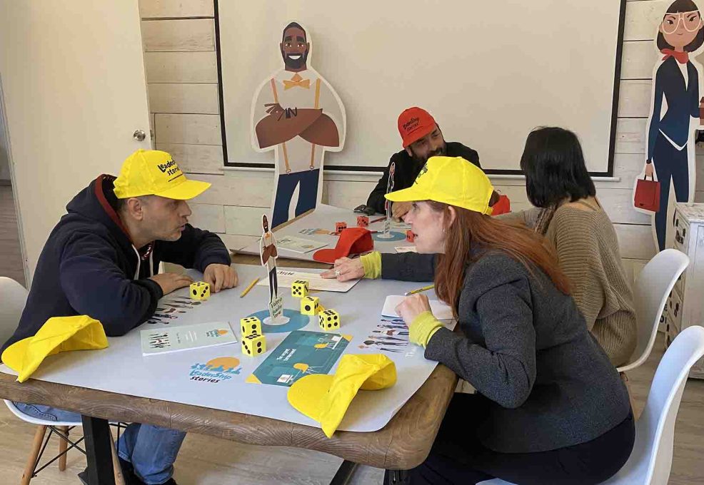 Team members engaging in the Leadership Stories game, wearing yellow hats and playing with game materials like dice and figurines, in a team building environment designed to enhance leadership skills.