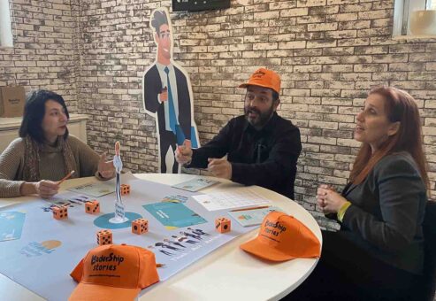 Participants engage in a Leadership Stories game, discussing and making leadership decisions, while wearing branded hats and working together at a table.
