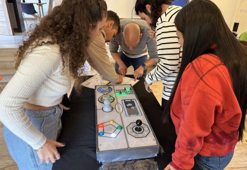Team members gather around the Team Synergy gaming console, collaborating and strategizing during a hands-on team building activity.