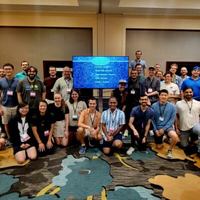 A large group of participants pose together in front of a Team Synergy retro gaming screen after a successful team building event, celebrating collaboration and teamwork. thumbnail