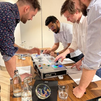 Team members collaborate around a Team Synergy retro gaming console, strategizing and engaging with controls to complete a mini-game challenge during a team building activity. thumbnail