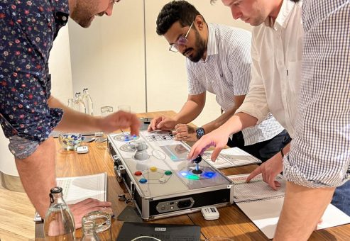 Team members collaborate around a Team Synergy retro gaming console, strategizing and engaging with controls to complete a mini-game challenge during a team building activity.