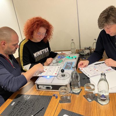 Three team members focus on the Team Synergy retro gaming console, analyzing instructions and working together to solve mini-game challenges during a team building event. thumbnail