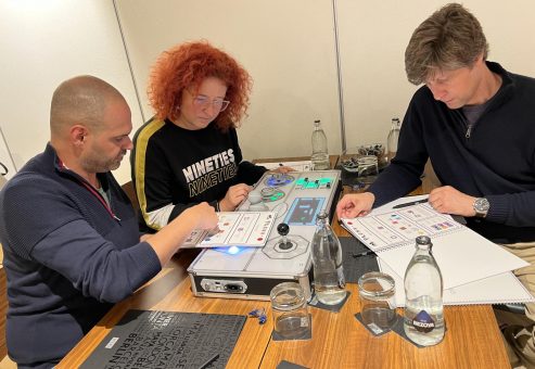 Three team members focus on the Team Synergy retro gaming console, analyzing instructions and working together to solve mini-game challenges during a team building event.