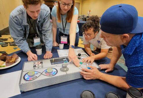 A group of participants works together on a Team Synergy gaming console, strategizing and solving challenges during a team building event in a conference room.